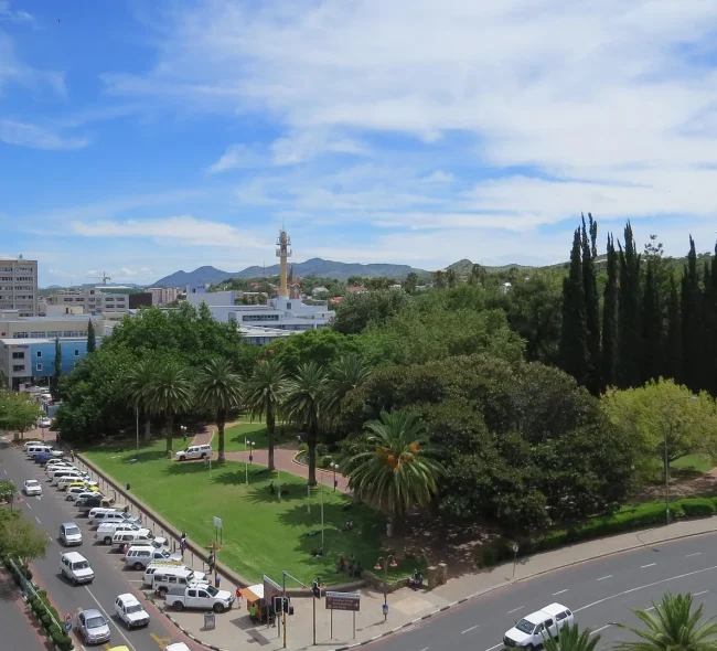 High-angle view of Windhoek city center in Namibia, featuring Zoo Park with lush green trees, palm-lined streets, and parked cars under a blue sky.